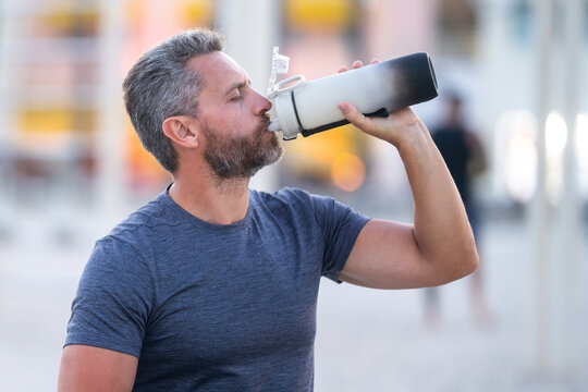 Sport fitness man resting on a bench and drink water from sport water bottle. Mature sport model sitting on a bench after a workout. Man taking break after fitness drink water from sport water bottle. - Powered by Adobe