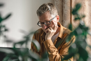 man sits at desk in his home, deep in thought with his hand on his chin. green plants frame scene, creating serene atmosphere for his focused work. close up.
