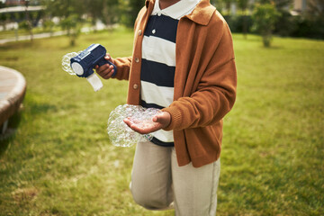 Black child holding bubble gun and catching bubbles in hand outdoors, wearing casual clothing, standing on grass in park, engaging in playful activity during daytime