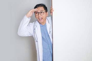 Joyful young male doctor with stethoscope peeking from behind blank white banner, ideal for...