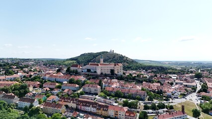 Obraz premium Aerial view of a historic castle surrounded by red-roofed houses and overlooked by a green hill with a chapel on top, capturing heritage and architectural beauty.