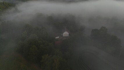 A solitary white house emerges through thick morning fog surrounded by dense trees, creating a...