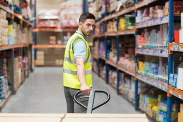 Man wearing safety vest pushing manual pallet jack along warehouse aisle with metal racks and boxes
