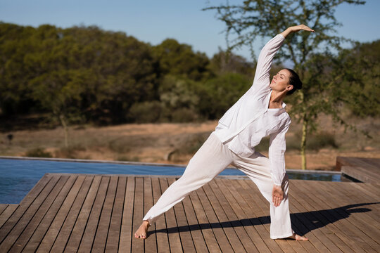 Woman wearing white yoga outfit performing side-stretch on wooden deck beside pool with trees