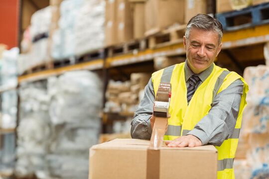 Male worker wearing vest using tape dispenser sealing cardboard box at packing station, copy space - Powered by Adobe