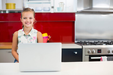Female child using laptop at kitchen island near plastic, ceramic bowls, pastel mugs, gas range