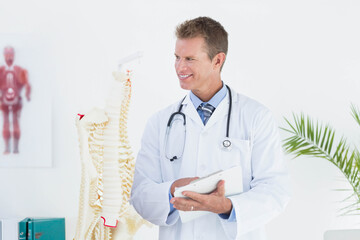 Middle-aged male physician holding clipboard beside spine model and wearing stethoscope in clinic