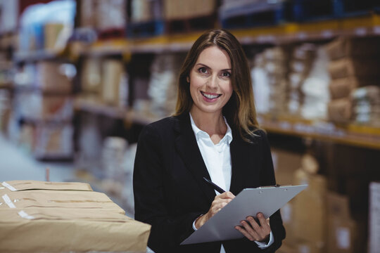 Businesswoman in black blazer writing on clipboard standing among warehouse shelves with boxes
