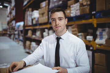 Male warehouse supervisor standing behind table reviewing forms in warehouse by shelves of boxes