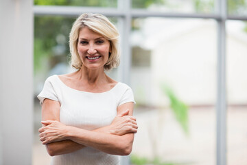 Senior woman standing wearing white off shoulder top by glass windows, showing green foliage