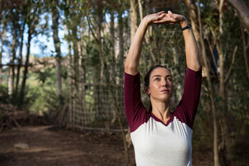 Woman stretching arms above head on forest trail wearing athletic shirt with fitness tracker