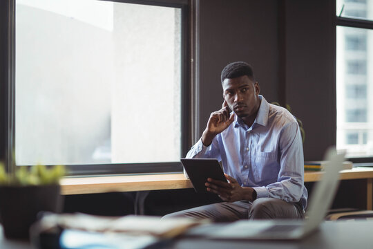 African American man sitting on bench by window in office holding tablet near laptop - Powered by Adobe