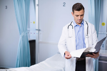 male doctor standing beside privacy curtains in cubicle reviewing file with stethoscope, copy space