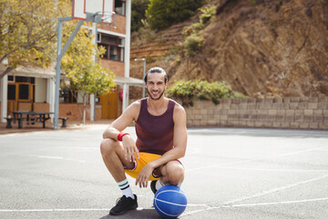 Adult man in sportswear kneeling at outdoor basketball court gripping ball near metal hoop
