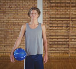 Teenage boy athlete smiling while holding blue basketball against brick wall in school gym
