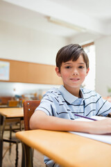 Fototapeta premium White boy student sitting at wooden desk in classroom, writing in open notebook with pencil
