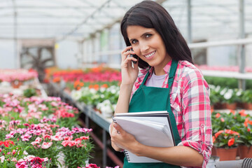 Woman greenhouse worker using notebook and talking on smartphone in potted plant aisles, copy space