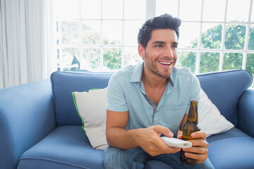 Man holding beer bottle, TV remote control sitting on blue sofa near window showing garden view