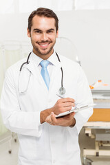 Male doctor wearing lab coat and blue tie holding clipboard in exam room with stethoscope