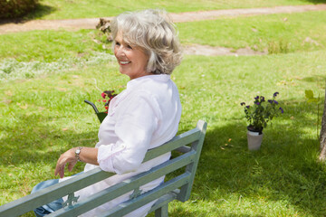 Senior woman sitting on bench in garden with wheelbarrow of red flowers and potted purple plants