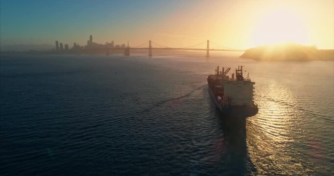 A container ship sails toward the San Francisco Bay Bridge at sunset, transporting goods. The city skyline is visible in the background, USA.