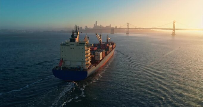 Cargo ship carrying containers sails into San Francisco Bay, USA, with the Bay Bridge and city skyline in the background at sunrise, for global trade.