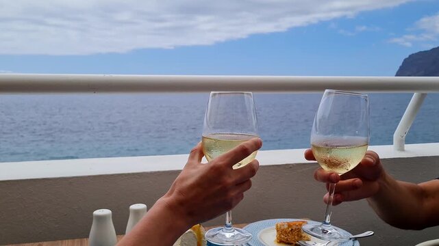 Romantic partners hold glasses of expensive wine and touch them while sitting in a restaurant on the ocean shore. Close-up. Young couple celebrating anniversary