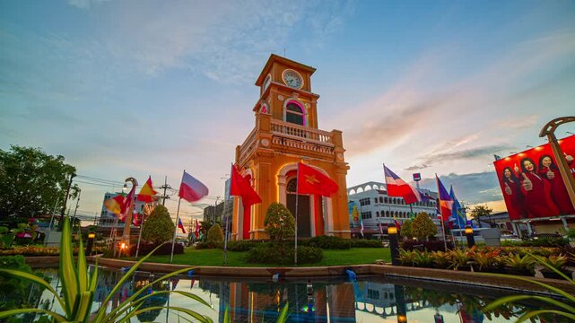 Phuket-Thailand, 13 August 2025: Time lapse A vibrant view of a historic yellow clock tower surrounded by flags and reflected in a pond under a dramatic sunset sky, capturing both cultural charm 