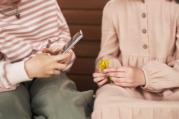 Two Caucasian children sitting close together, one child holding smartphone and using camera while other child holding small bouquet of yellow wildflowers in hands