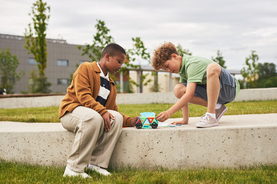 Two boys, one Black and one Caucasian, sitting on concrete bench outdoors, interacting with educational robot toy and cards, focused on collaborative learning activity
