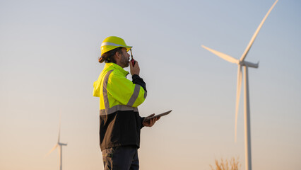 Engineer working on laptop in front of wind turbines for checking wind turbines of the field during beautiful sunset. Alternative energy concept.