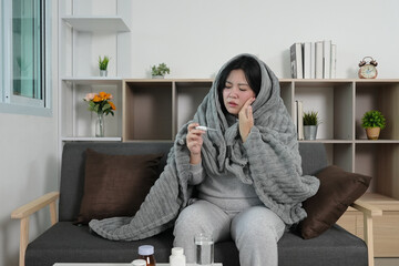 Health and Wellness. Woman wrapped in a blanket, checking her temperature at home.