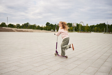 Caucasian girl riding scooter outdoors on spacious paved area, long hair flowing behind, wearing...