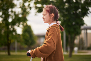 Caucasian girl riding scooter outdoors, looking forward with focused expression, standing in park...