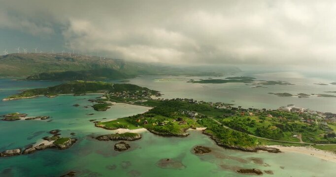 Drone pullback reveals Sommaroy islands and turquoise shallows near Troms&oslash;, with a distant ridge-top wind farm and a broad fjord horizon under soft summer clouds.