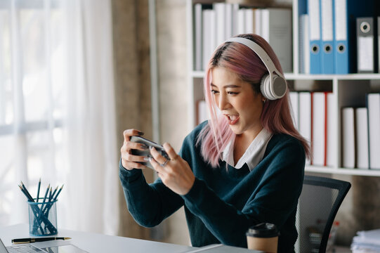Joyful young gamer girl with headphones playing mobile game at office desk. Real people enjoying digital lifestyle, online entertainment, and casual gaming fun. - Powered by Adobe