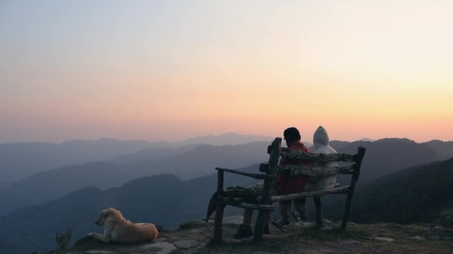 Two men sitting on a bench chairs enjoy sunset and chatting outdoors at the Top of Raghupur Fort - Tirthan Valley Himanchal pradesh india.	