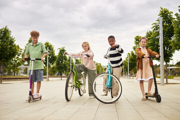 Group of four children including two Caucasian girls, one Caucasian boy, and one Black boy riding scooters and bicycle outdoors on paved walkway