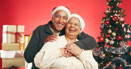 Hug, Christmas and portrait of senior couple in studio with love, festive and marriage tradition. Smile, santa hat and elderly man embracing woman by xmas tree for celebration on red background.