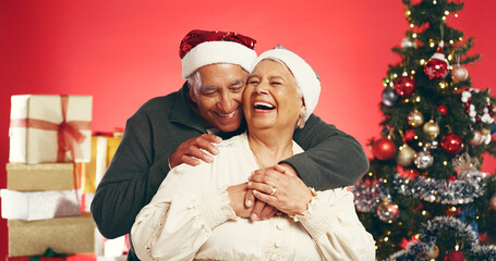 Laugh, christmas and senior couple in studio with love, festive and marriage tradition. Hugging, santa hat and elderly man embracing woman by xmas tree for celebration together by red background.