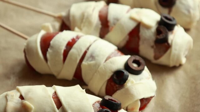 raw Halloween appetizer mummies made from sausages wrapped in puff pastry and decorated with olives. Festive spooky snack idea, ready to be baked for Halloween party food