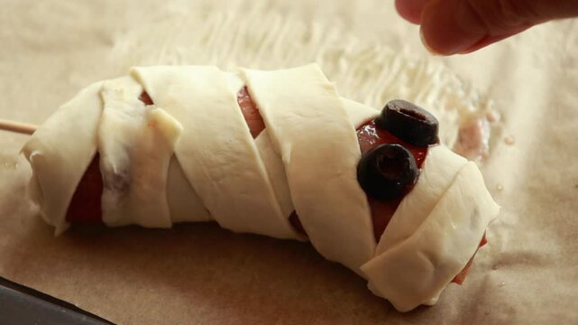 raw Halloween appetizer mummies made from sausages wrapped in puff pastry and decorated with olives. Festive spooky snack idea, ready to be baked for Halloween party food
