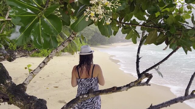 Young woman with white hat and zebra striped dress walking on a beautiful tropical beach under tree branches, enjoying her vacation in a rainy nasty day