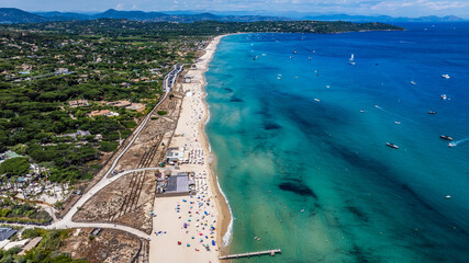 aerial view along Pampelonne Beach landscape to Saint-Tropez - located at coastline of Ramatuelle, Côte d’Azur, France - with people on the beach in water, green wooded hilly landscape in background