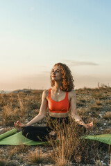 A woman practices yoga in a lotus position on a hilltop at sunset