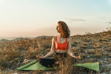 A woman practices yoga in a lotus position on a hilltop at sunset