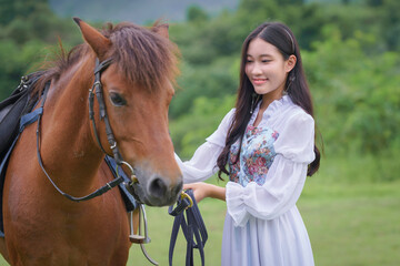 Pretty young Asian woman traveler in a beautiful modern dress enjoys horse training at a natural outdoor horse riding field.