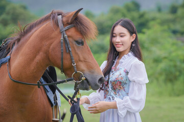 Obraz premium Pretty young Asian woman traveler in a beautiful modern dress enjoys horse training at a natural outdoor horse riding field.