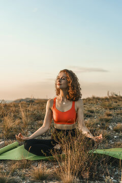 A woman practices yoga in a lotus position on a hilltop at sunset