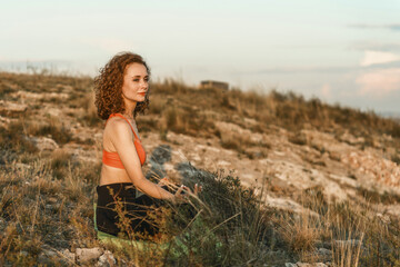 A woman practices yoga in a lotus position on a hilltop at sunset